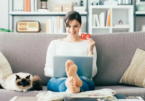 Young smiling woman at home, she is relaxing on the couch with her cat and shopping online using a credit card